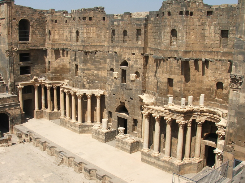 Les comédiens se produisaient sur l’étroit pulpitum au pied d’un monumental mur de scène (frons scaenae) en partie curviligne. À partir de colonnes corinthiennes intactes, l’ordre du niveau inférieur a été partiellement restitué. Il faut imaginer ce décor somptueux qui date de l’époque sévérienne (193-235 ap. J.-C.), sur trois étages. Les niches étaient ornées de statues. Le mur qui contribuait à la qualité acoustique est percé de trois portes par lesquelles entraient et sortaient les acteurs.
CC 4.0 Odysseum, photo F. Poli
Articles à consulter :
L’architecture du théâtre - Chronologie de l’histoire romaine - Glossaire du musée virtuel
Bosra, frons scaenae (mur de scène) du théâtre .JPG