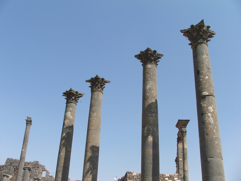 Au carrefour sud des deux principaux axes de la ville s’élèvent quatre colonnes aux chapiteaux corinthiens. Elles ornaient la façade d’un monument public dressé de hauts murs avec une grande abside centrale. À l’arrière-plan, à droite, deux colonnes coiffées d’un entablement richement décoré sont les vestiges probables d'un nymphée, fontaine monumentale. La ville construite sur un plateau basaltique a fourni le matériau de ces édifices qui ont résisté à l’érosion et aux tremblements de terre.
CC 4.0 Odysseum, photo F. Poli
Article à consulter :
Glossaire du musée virtuel
Bosra, l’exèdre monumentale et le nymphée.JPG