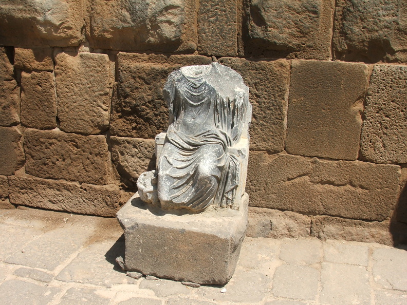 On ignore si cette statue de Sérapis ornait les thermes ou appartenait à un édifice cultuel disparu. Sculptée dans un schiste égyptien, la statue est une pièce d’importation. Le dieu, vêtu d’une tunique (chiton) longue et d’un ample manteau (himation) est représenté assis sur un trône à haut dossier et accoudoirs. À ses pieds se tient un animal à trois têtes, aujourd’hui cassées, sans doute le chien Cerbère, gardien de l’entrée des Enfers. Sérapis était souvent assimilé au dieu chtonien Hadès.
CC 4.0 Odysseum, photo F. Poli
Articles à consulter :
Dieux grecs et romains - Glossaire du musée virtuel
Bosra, statue de Sérapis, thermes du centre.JPG