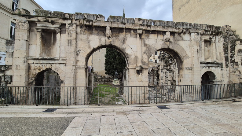 L’une des plus longues enceintes fortifiées de la Gaule romaine protégeait Nemausus. Construite en 16 av. J.-C., la Porte Auguste était implantée sur la Via Domitia qui reliait l’Italie à la péninsule ibérique. Flanquée de deux tours défensives, elle atteint près de 40 mètres de long. Les véhicules empruntaient les grands arcs centraux et les deux petits arcs latéraux étaient destinés aux piétons. Le monument s'ouvrait sur une vaste cour bordée de portiques, dans le prolongement des arcades.
Photos CC 4.0 Odysseum, classe de P. Cochet Terrasson 2025
Articles à consulter :
Auguste - Glossaire du musée virtuel
Nîmes, la Porte Auguste.jpg